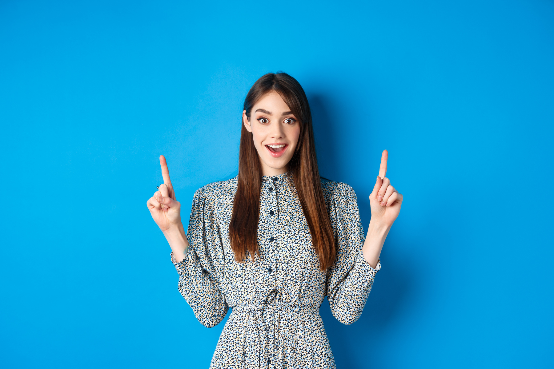 Excited and Happy Young Woman in Dress Checking Out Special Offer, Pointing Fingers up and Smiling, Standing against Blue Background